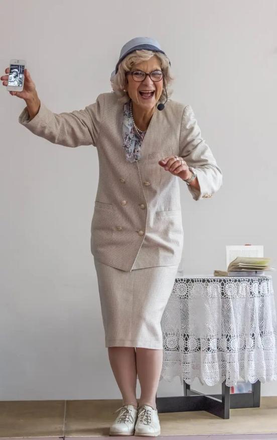 Die Schauspielerin Anke Siefken als die Seniorin Reanta Bergmann, im beigen Kostüm und mit grauer Perücke. Sie hält ein Handy hoch