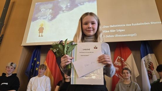 Vika Eliv Löper hält eine Urkunde, einen Blumenstrauß und einen Briefumschlag in den Händen.